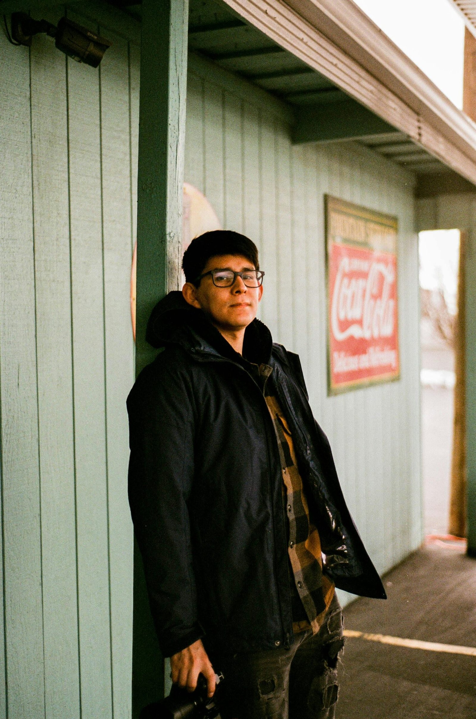 Man in glasses and jacket standing on a wooden porch with a vintage sign.